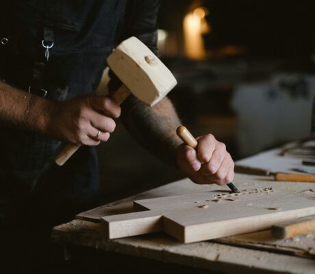 Unrecognizable male artisan using wooden hammer and chisel to carve lumber plank while working at table with special instruments on blurred background