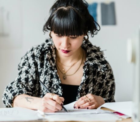 Concentrated Asian female designer with red lipstick drawing draft while sitting at table with sewing machine in modern light workshop